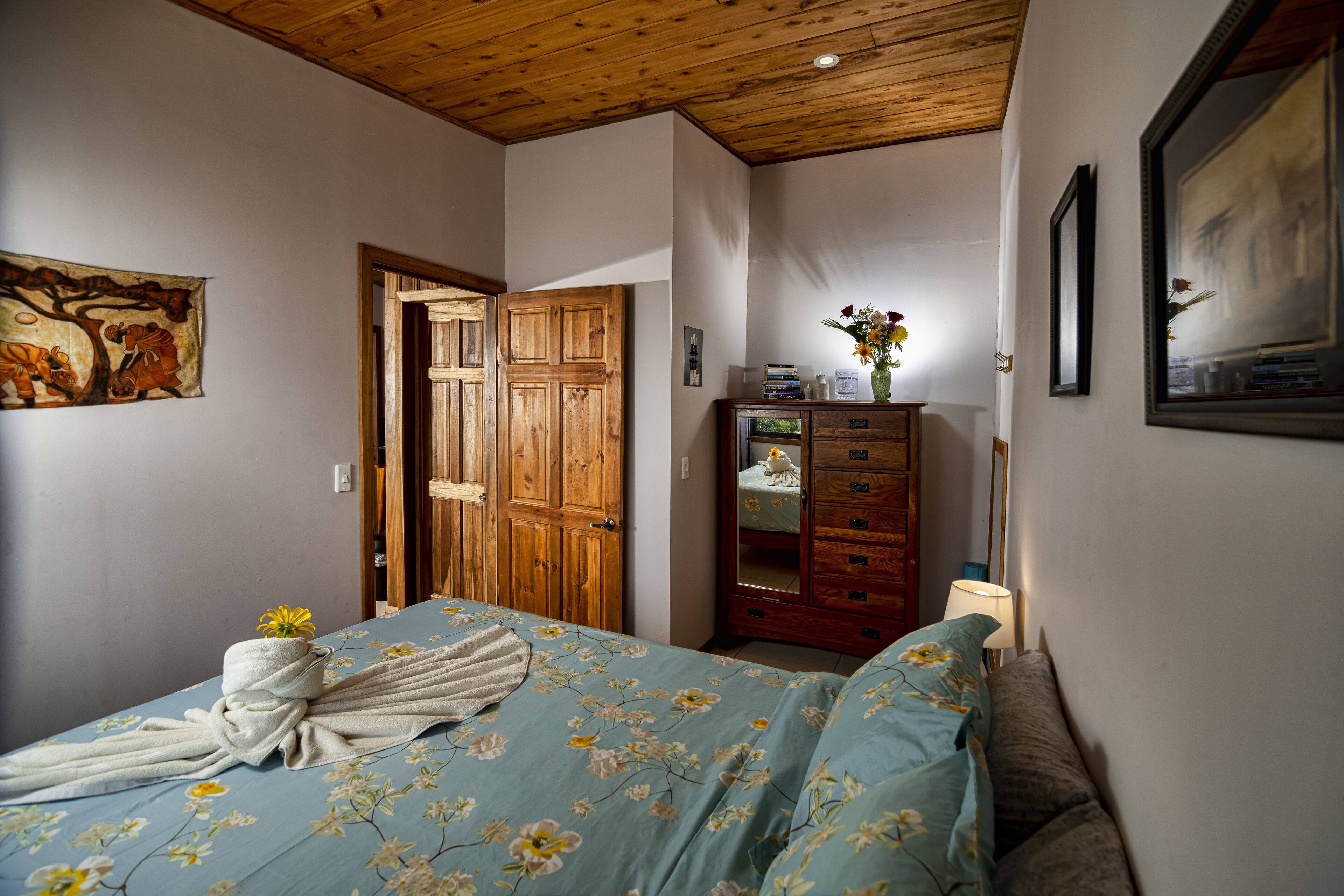 A bedroom with a wooden ceiling, a bed with floral bedding, a wooden wardrobe, and a painting on the wall.