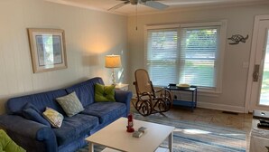 A living room with a blue sofa, a white coffee table, a wicker chair, and a window with blinds. - Seaside Serenity, Steps to Beach, Pet Friendly (Folly Beach)