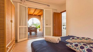 Une chambre à coucher comprenant un lit, une armoire en bois et une porte-fenêtre coulissante donnant sur une terrasse extérieure avec une table et des chaises.