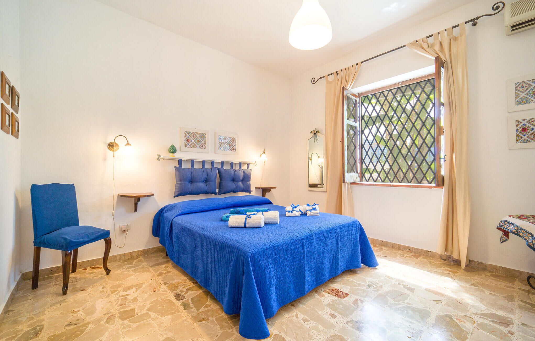 A bedroom with a bed, a chair, a nightstand, a window with curtains, and a wall-mounted air conditioner.