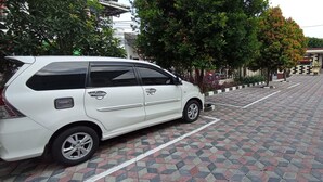 A white minivan parked on a patterned pavement with trees and buildings in the background. - Wisma Anton Soedjarwo (Jakarta)