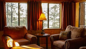 Una habitación acogedora con un sofá de cuero, un sillón a juego y una mesita de noche con una lámpara. Ventanas amplias con cortinas ofrecen vistas a árboles al exterior.