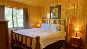 Un dormitorio de cabaña de madera con una cama, dos lámparas de noche, un ventanal y un cuadro en la pared.