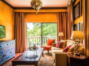 A room with a sofa, a coffee table, a lamp, a TV, and a view of greenery outside. - Avani Victoria Falls Resort (Livingstone)