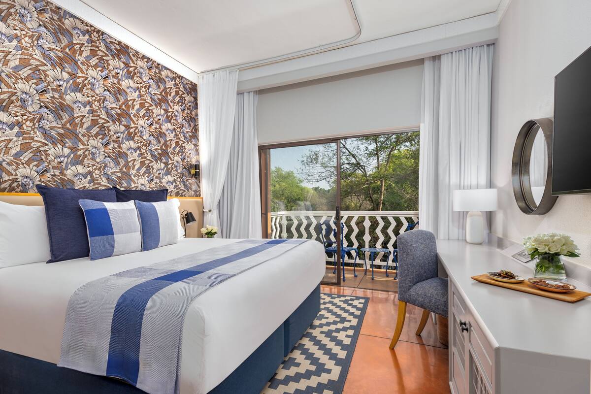 A modern hotel room with a large bed, a desk, a chair, and a balcony with a view of trees.