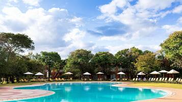 Piscina con sillas de descanso y sombrillas, rodeada de árboles y un cielo despejado.