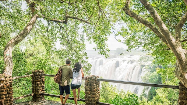 A couple standing on a stone bridge overlooking a waterfall, surrounded by lush greenery. - Avani Victoria Falls Resort (Livingstone)