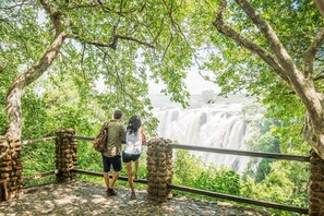 A couple standing on a stone bridge overlooking a waterfall, surrounded by lush greenery. - Avani Victoria Falls Resort (Livingstone)