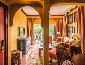 Una habitación con sofá, sillones, televisor, alfombra estampada y balcón con cortinas.