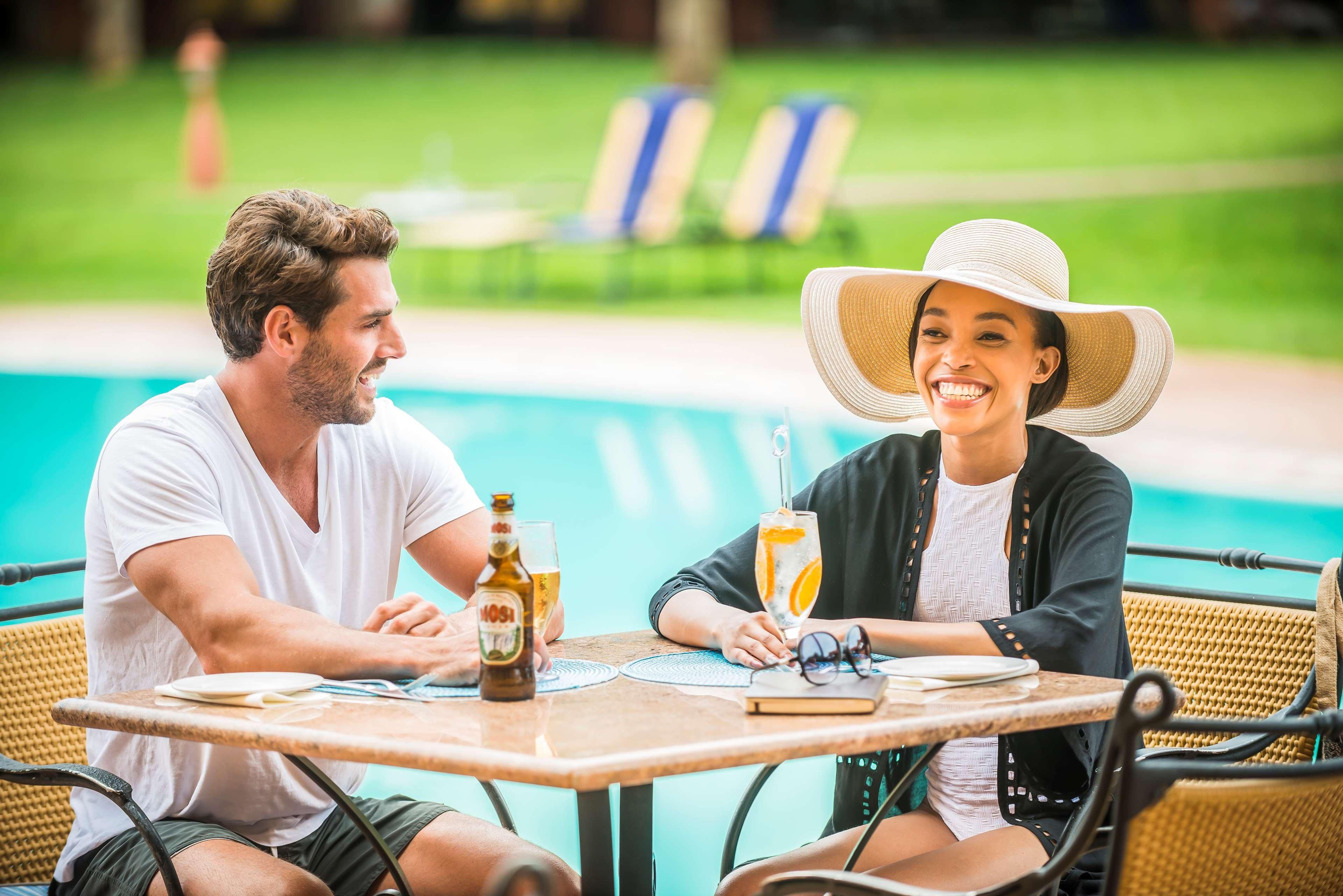 Duas pessoas sentadas à mesa perto de uma piscina, com uma garrafa de cerveja e um copo de suco de laranja sobre a mesa.