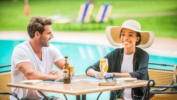 Duas pessoas sentadas à mesa perto de uma piscina, com uma garrafa de cerveja e um copo de suco de laranja sobre a mesa.