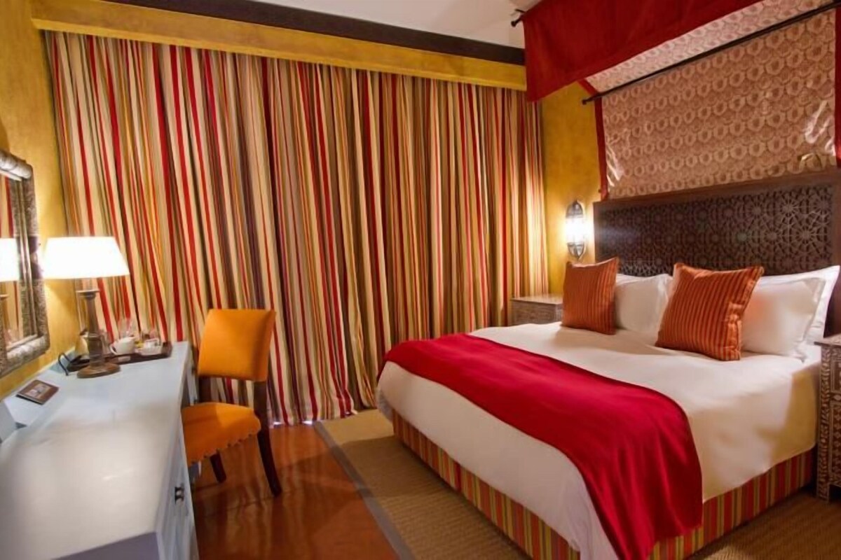 A hotel room with a bed, a desk, a chair, and a patterned nightstand.