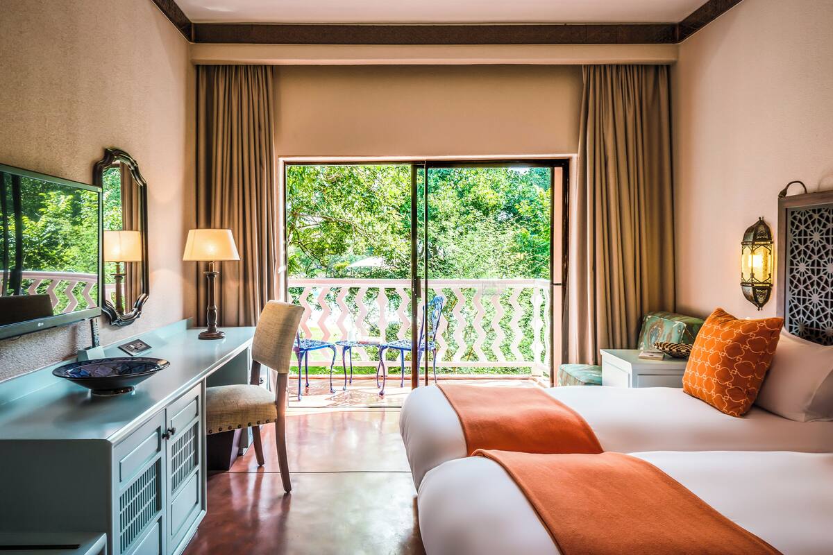 A hotel room with a bed, a desk, a chair, a balcony with a view of greenery, and a window with curtains.