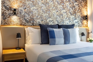 A hotel room with a patterned wallpaper, a neatly made bed with blue and white bedding, and two bedside lamps. - Avani Victoria Falls Resort (Livingstone)