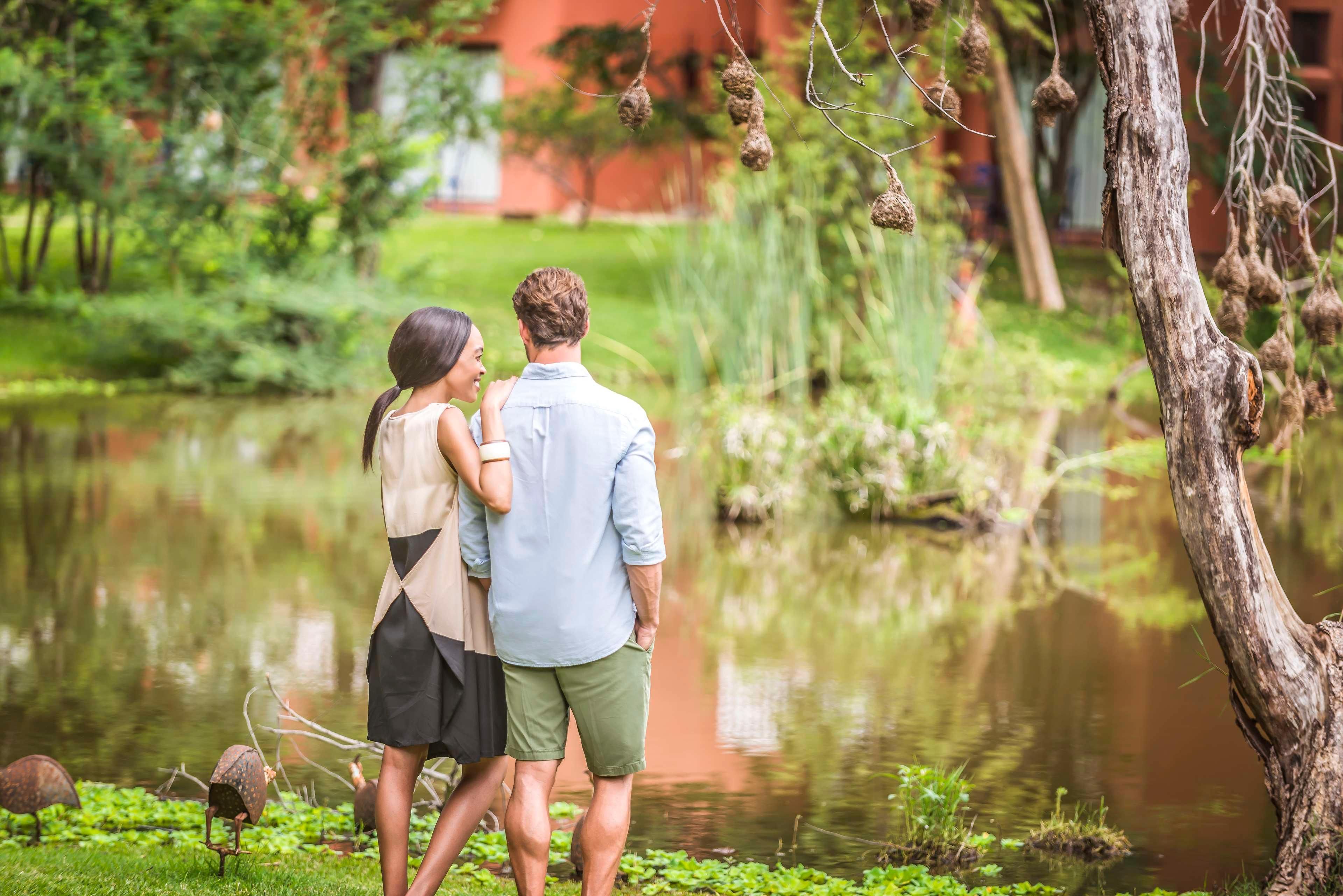 Un couple se tient près d’un étang, avec de la verdure et un arbre en arrière-plan.