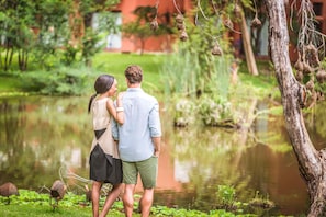 Una pareja parada junto a un estanque, con follaje y un árbol de fondo.