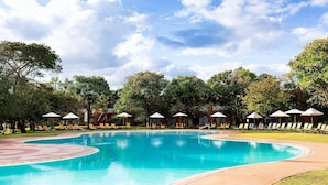A swimming pool with lounge chairs and umbrellas, surrounded by trees and a clear sky. - Avani Victoria Falls Resort (Livingstone)
