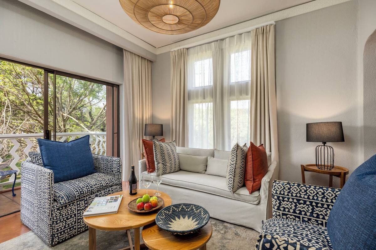 A modern living room with patterned furniture, a dining table with fruit, and a view of trees through large windows.