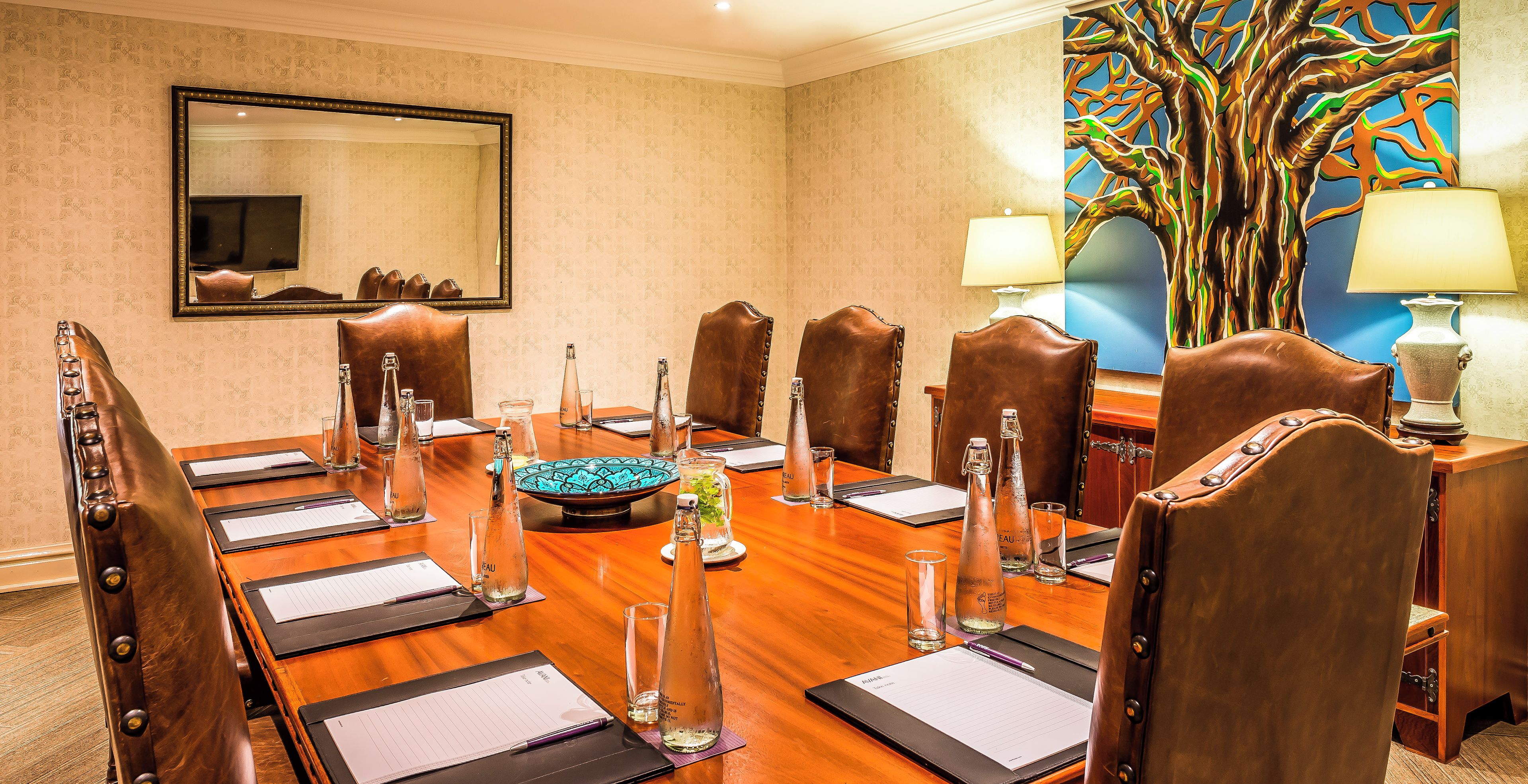 A conference room with a long table, leather chairs, a large mirror, and a colorful tree painting.