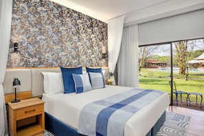 A modern bedroom with a large bed, a nightstand, a wall with a floral pattern, and a view of a garden through a large window. - Avani Victoria Falls Resort (Livingstone)