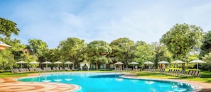 A swimming pool with lounge chairs and umbrellas, surrounded by trees. - Avani Victoria Falls Resort (Livingstone)