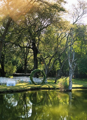 Un salón de bodas al aire libre con filas de sillas y mesas blancas, rodeado de árboles y una masa de agua.