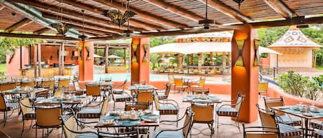 Un espacio exterior con mesas y sillas para comer, una piscina al fondo y una estructura cubierta con techo estampado.