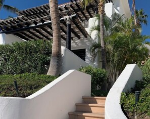 Un edificio blanco con un espacio exterior cubierto, palmeras y una escalera que conduce a la entrada.