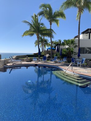 A swimming pool with lounge chairs and umbrellas, surrounded by palm trees and a view of the ocean. - CABO OCEANVIEW, WALK TO THE BEACH, CREATE MEMORIES  (San José del Cabo)