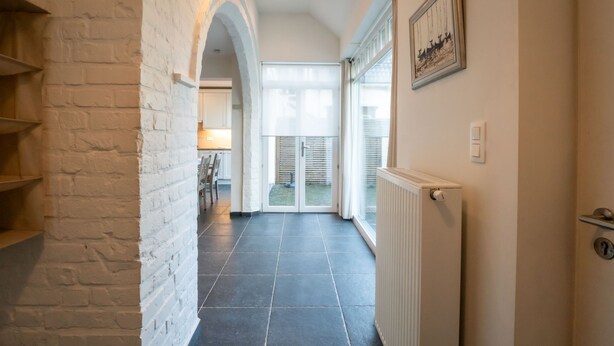 Un couloir avec un mur en briques blanches, un sol carrelé et un radiateur. Une porte donne sur une pièce avec une fenêtre et un tableau encadré au mur.