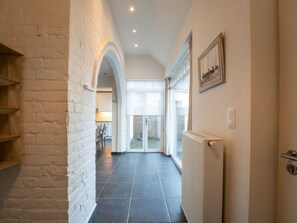 Un couloir avec un mur en briques blanches, un sol carrelé et un radiateur. Une porte donne sur une pièce avec une fenêtre et un tableau encadré au mur.