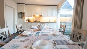 Un coin repas avec une table dressée pour quatre personnes, des meubles blancs et, au fond, une cuisine. Une grande fenêtre est agrémentée de rideaux.