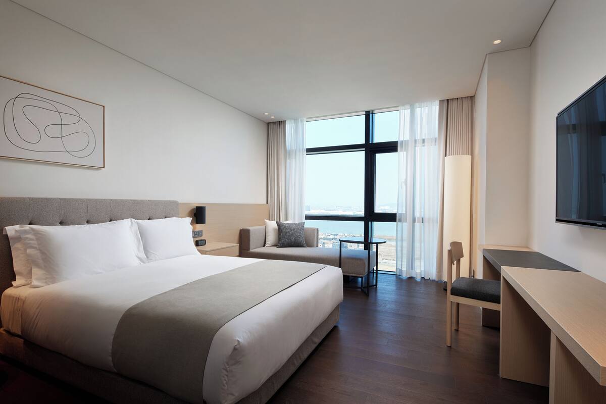Quarto de hotel moderno com uma cama grande, uma escrivaninha e vista para o mar através de amplas janelas.