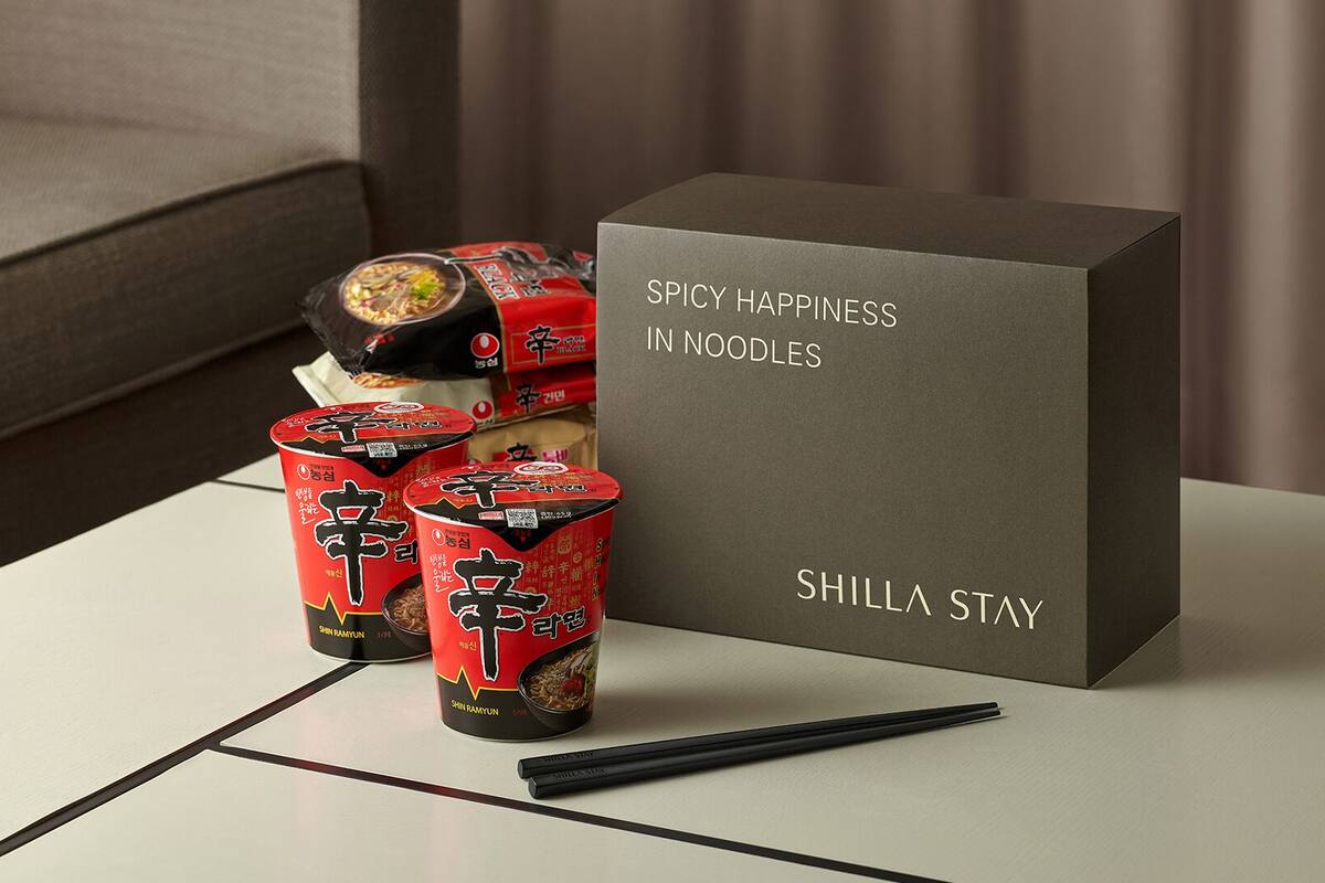 Uma mesa com uma caixa identificada como “FELICIDADE PICANTE EM MACARRÃO” e vários itens embalados em vermelho e preto, provavelmente comida, com hashis ao lado.
