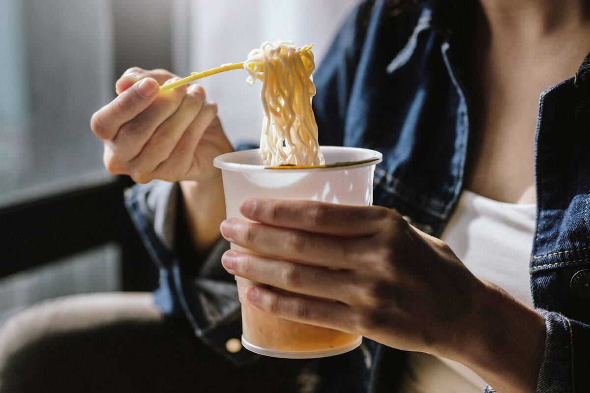 Uma pessoa segurando uma caneca com macarrão e uma colher amarela.