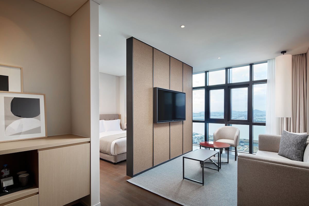 Quarto de hotel moderno com cama, sofá, televisão e uma grande janela com vista para a cidade.