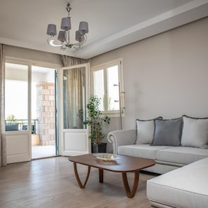 Un salon avec un canapé, une table basse, un lustre et une vue sur l’extérieur, visible à travers une porte vitrée.