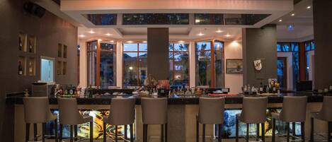 A bar area with multiple stools, a bar counter, and a large window with a view of the night sky.