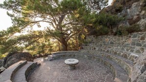 Un espacio circular de piedra con asientos y una mesa de piedra en el centro, rodeado de escalones y un muro de piedra.