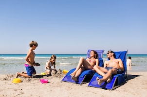 Escena de playa con gente descansando en tumbonas y un niño jugando con juguetes de arena.