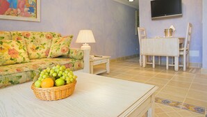 A living room with a floral sofa, a table with a fruit basket, a lamp, a TV mounted on the wall, and a dining area with a table and chairs. - Royal Son Bou Family Club (Alayor)