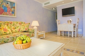 A living room with a floral sofa, a table with a fruit basket, a lamp, a TV mounted on the wall, and a dining area with a table and chairs. - Royal Son Bou Family Club (Alayor)