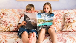 Two children sitting on a patterned sofa, reading a book titled 'Menorca'. - Club Royal Son Bou (Alayor)