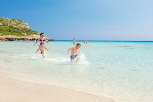Escena de playa con agua turquesa cristalina, orilla arenosa y acantilados rocosos al fondo.