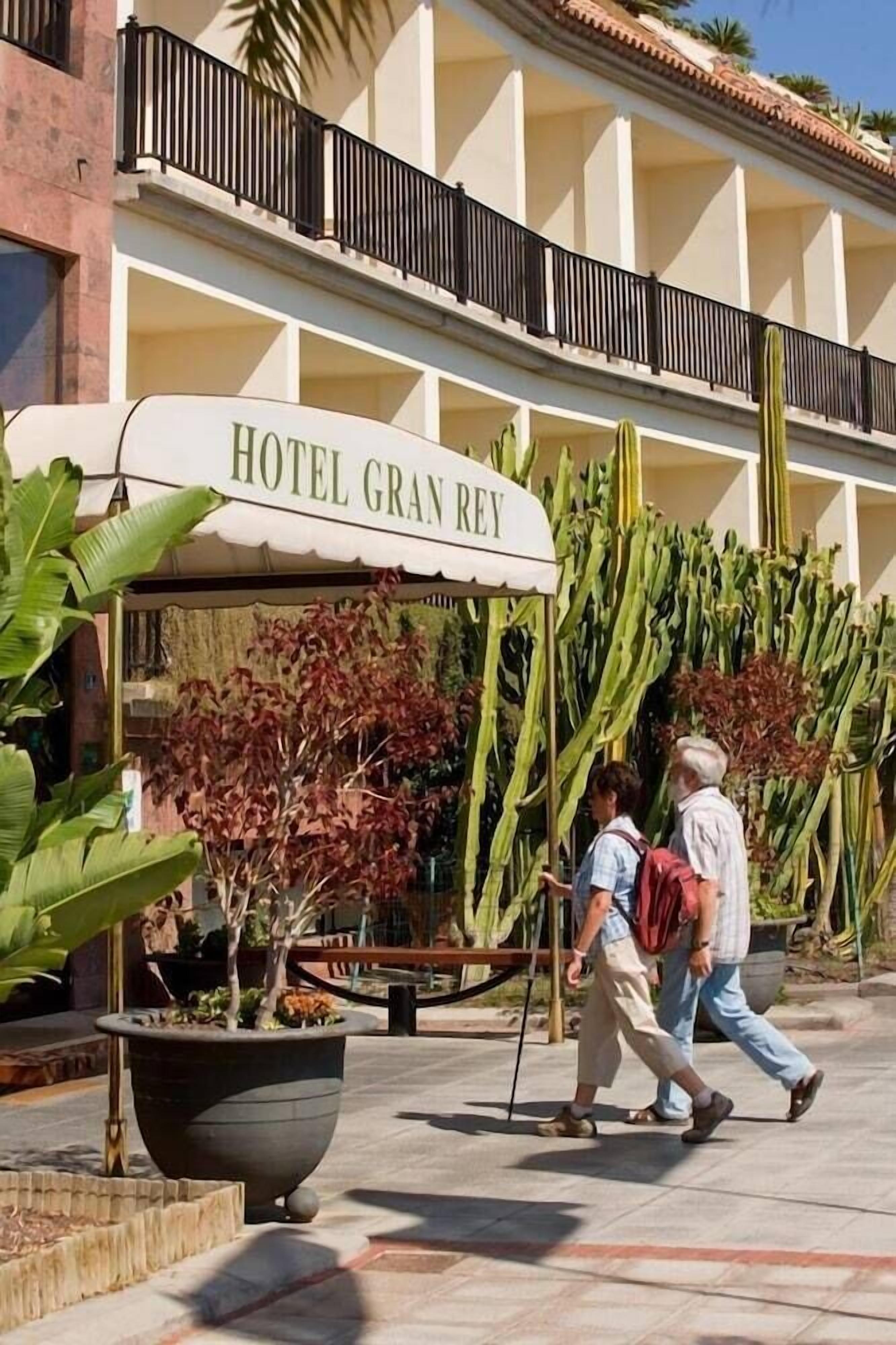 A hotel entrance with a sign reading "HOTEL GRAN REY," a large planter with plants, and a pedestrian walkway.