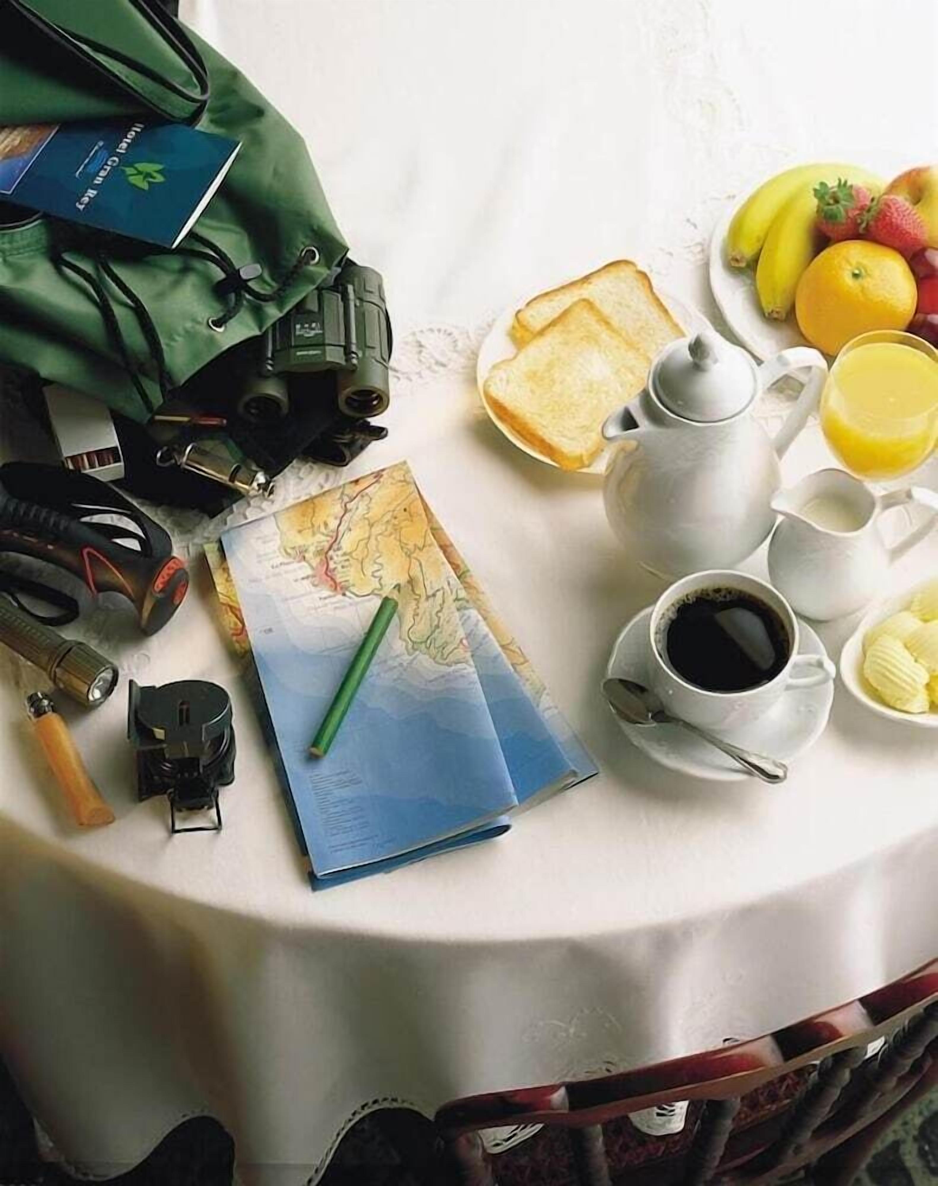 A table with a map, a green bag, a flashlight, a compass, a pair of binoculars, a cup of coffee, a teapot, a plate with bread, a banana, and a glass of orange juice.