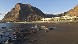 A beach with rocky shoreline, a mountain in the background, and a row of white buildings with balconies. - Gran Rey (Valle Gran Rey)