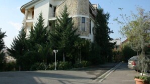 A two-story house with a stone tower, surrounded by evergreen trees and a parked car. - Baron (Tirana)