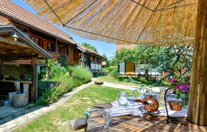 Uma área de refeições rústica ao ar livre, com uma mesa de madeira posta para dois, rodeada por vegetação exuberante e um guarda-sol de palha.