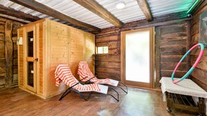 Une cabane en bois avec deux fauteuils de jardin à rayures, un sauna en bois et un cerceau de hula hoop posé sur une table.
